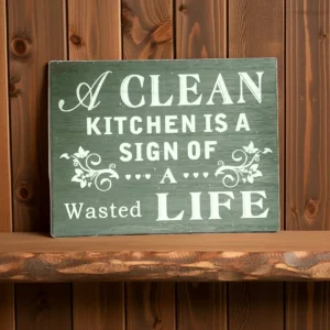 green wooden plaque with white text reading “A Clean Kitchen Is a Sign of a Wasted Life,” displayed on a wooden shelf.