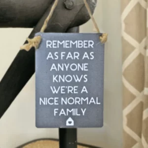 Blue mini metal sign with white text reading “Remember as far as anyone knows we’re a nice normal family,” hanging by jute string.