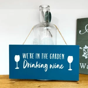 Blue rectangular hanging sign with white text reading “We’re in the garden drinking wine”, decorated with wine glass icons and hanging by a jute rope.