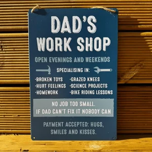 Navy blue metal hanging sign reading “Dad’s Work Shop – Open Evenings and Weekends. If Dad Can’t Fix It Nobody Can”, displayed on a wooden wall.