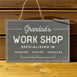 Grey rectangular wooden sign reading “Grandad’s Work Shop – Specialising in Mending, Making, Miracles”, hanging on a wooden wall with jute string.
