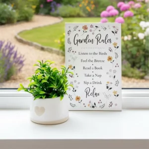 White hanging sign with floral border and text listing garden rules: “Listen to the Birds, Feel the Breeze, Read a Book, Take a Nap, Sip a Drink, Relax”, displayed beside a potted plant.