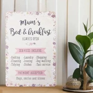 Pastel floral metal sign reading “Mum’s Bed & Breakfast – Always Open”, listing humorous household services and “payment accepted: hugs, smiles and kisses”. Displayed on a light wooden surface beside a green houseplant.