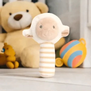 Soft beige and cream lamb cuddle stick rattle standing upright on a nursery floor with toys and a teddy bear in the background.
