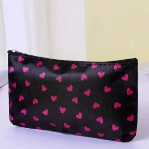 Small black cosmetic bag with pink heart pattern and silver zip pull, displayed on a white table beside beauty accessories.