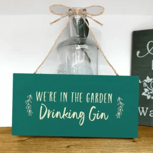Green wooden hanging sign with the text “We’re in the garden drinking gin”, hung by a jute rope.